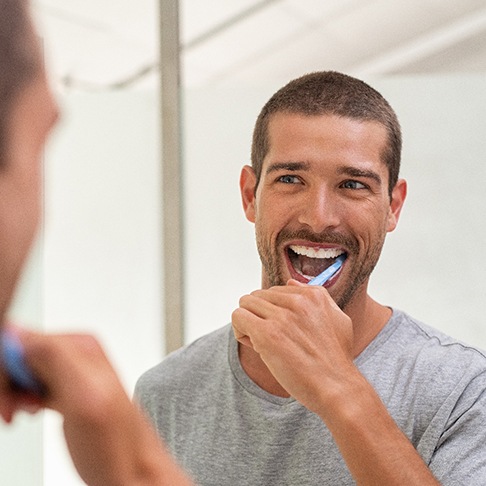 A smiling man brushing his teeth in front of a bathroom mirror